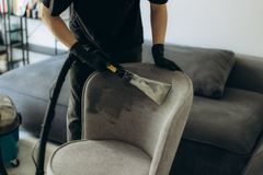Person wearing black gloves vacuums a gray upholstered chair with a cleaning tool in a living room.