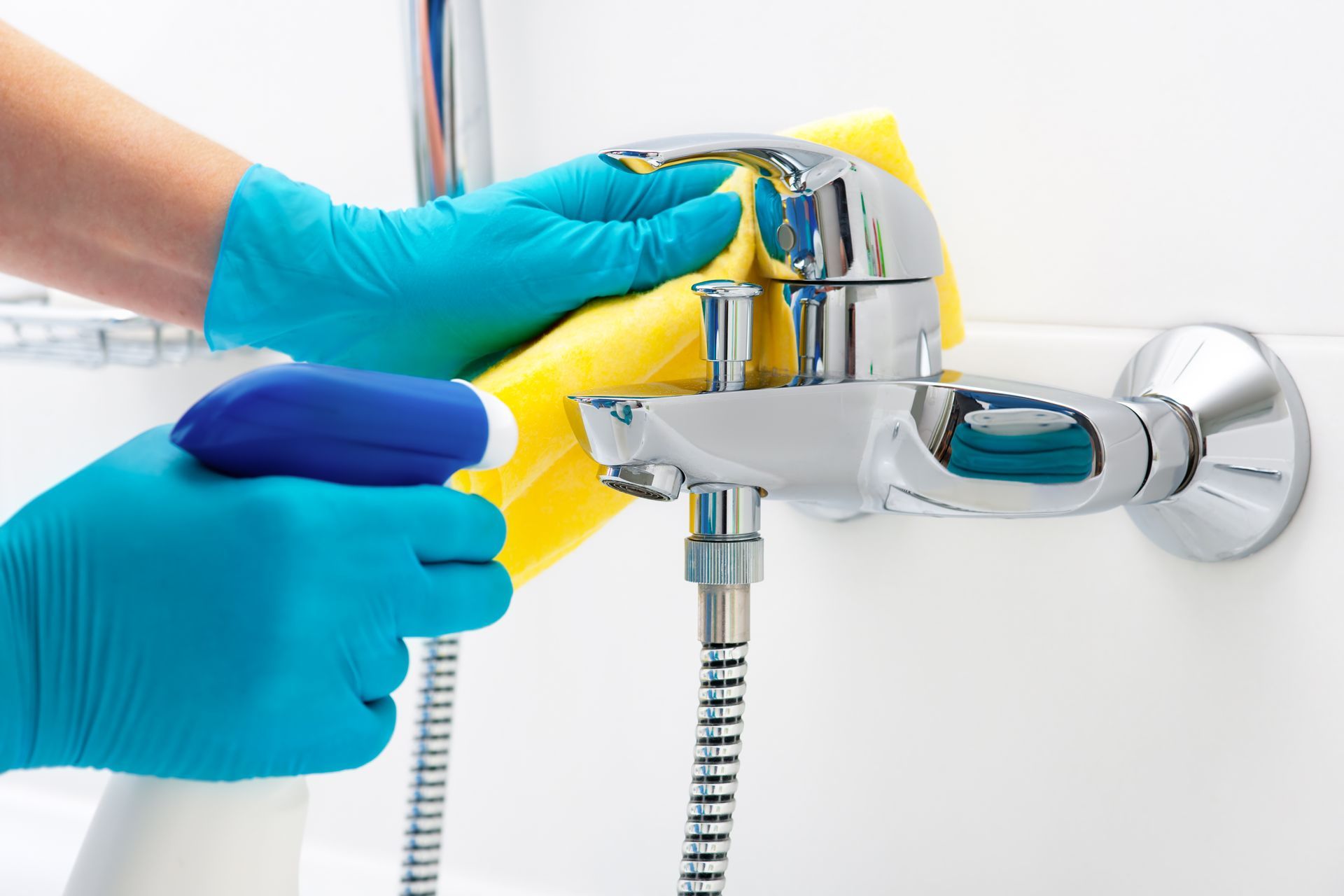 Person wearing blue gloves sprays and wipes a chrome faucet in a white bathroom.