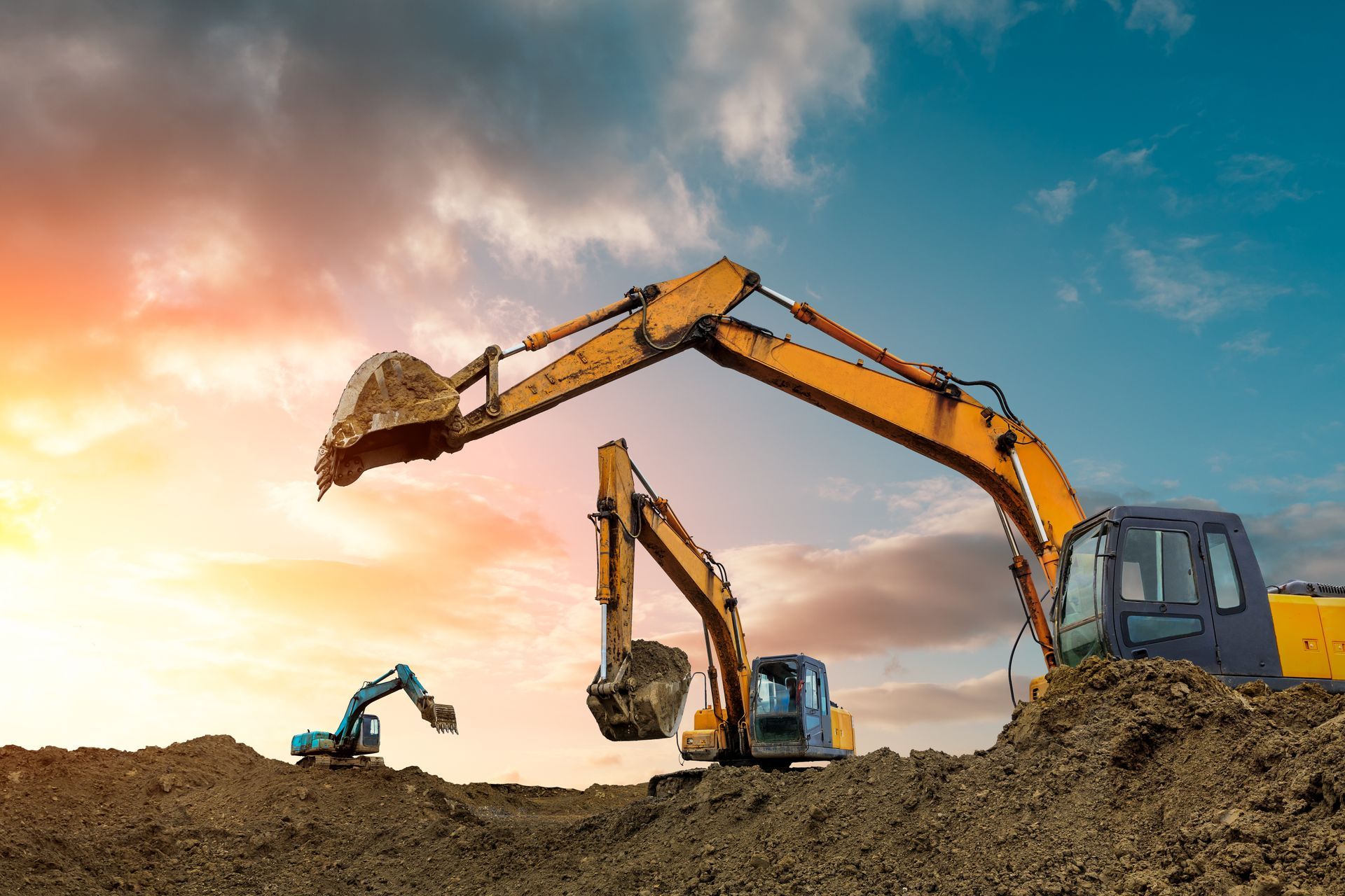 Three yellow and blue excavators working on a dirt mound under a sunset sky.