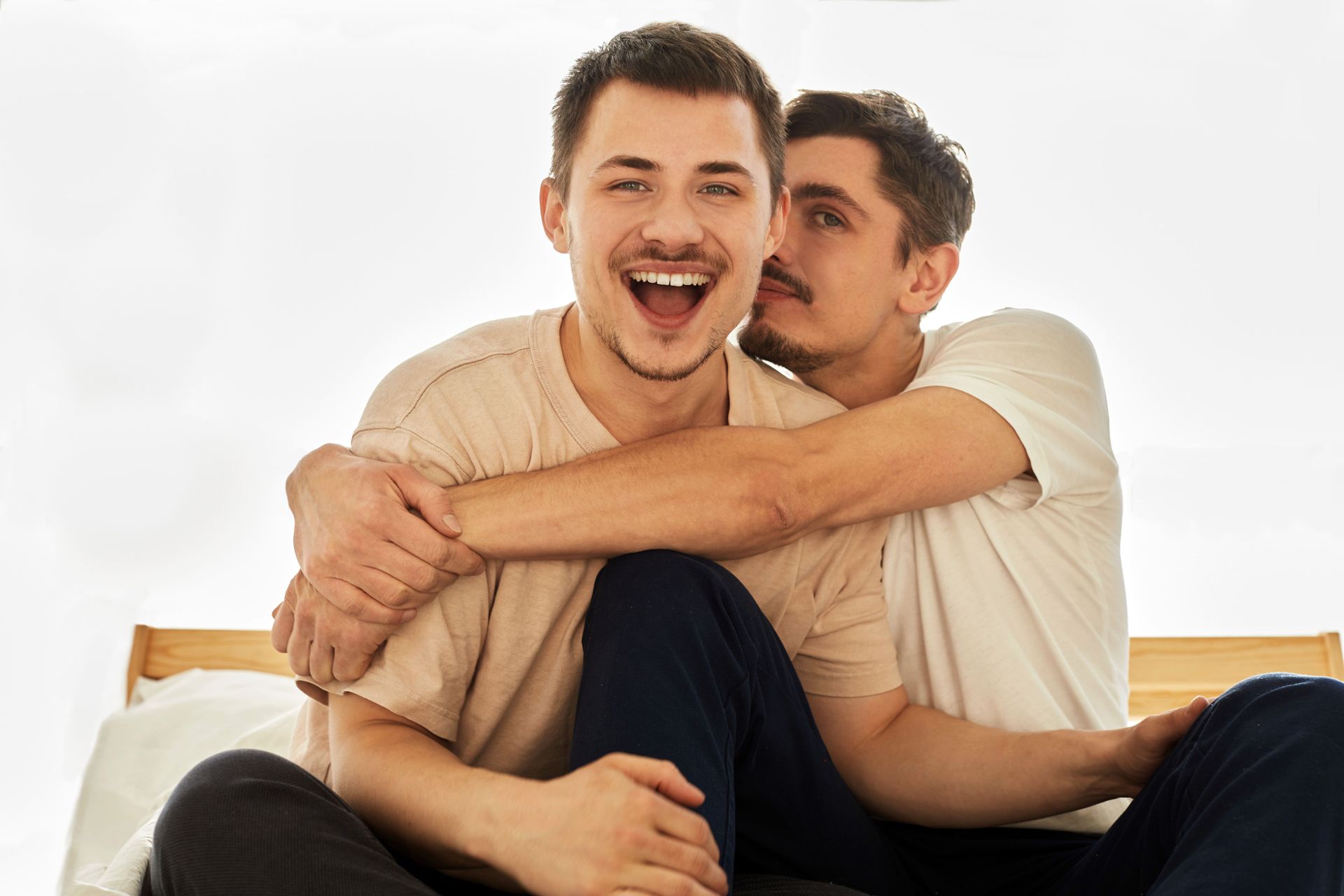 Two men are sitting on a bed, hugging each other.