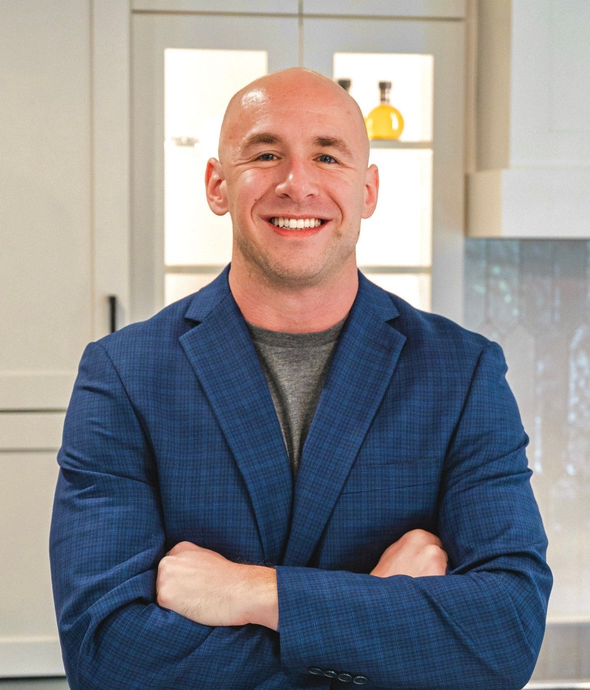 Bald man in blue blazer smiles, arms crossed, in a kitchen.