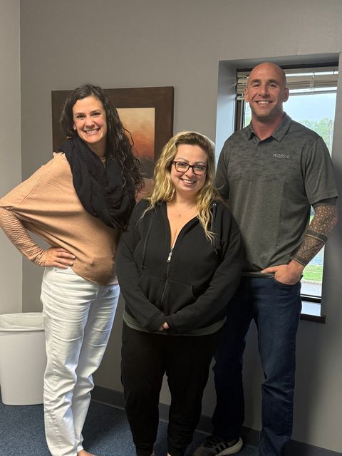Three people smiling in an office. Woman with curly hair and scarf, a woman in glasses, and a man with tattoos.
