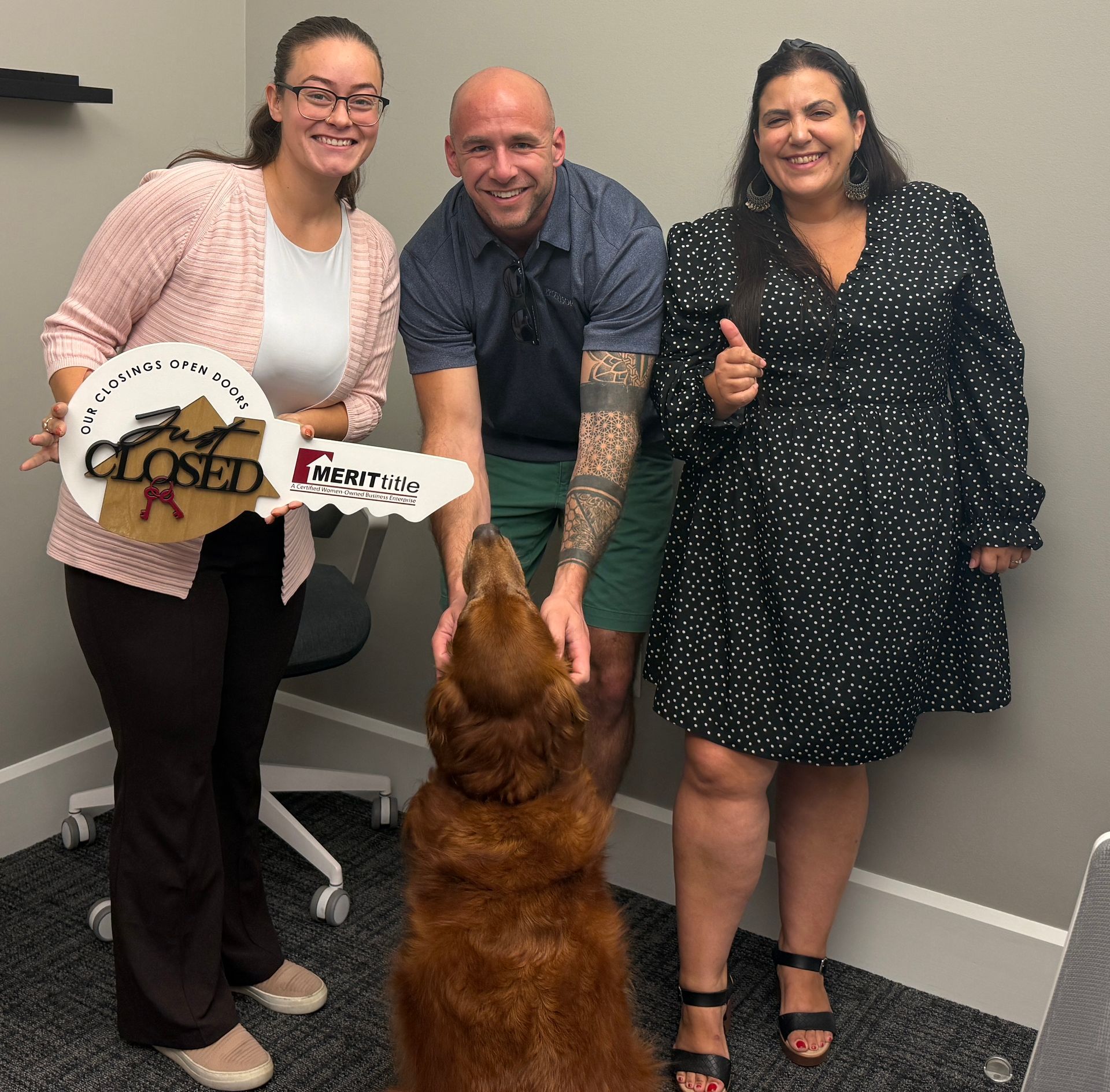 Three people and a dog celebrate a home closing, holding a large key in an office.