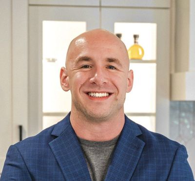 Bald man smiling, wearing a blue blazer and gray shirt, standing in a kitchen.