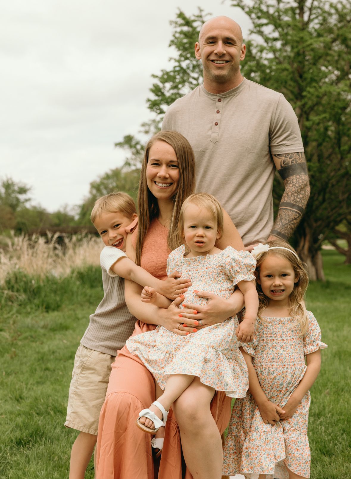 Family of five posing outdoors: woman holding toddler, other children flanking her, man standing behind.