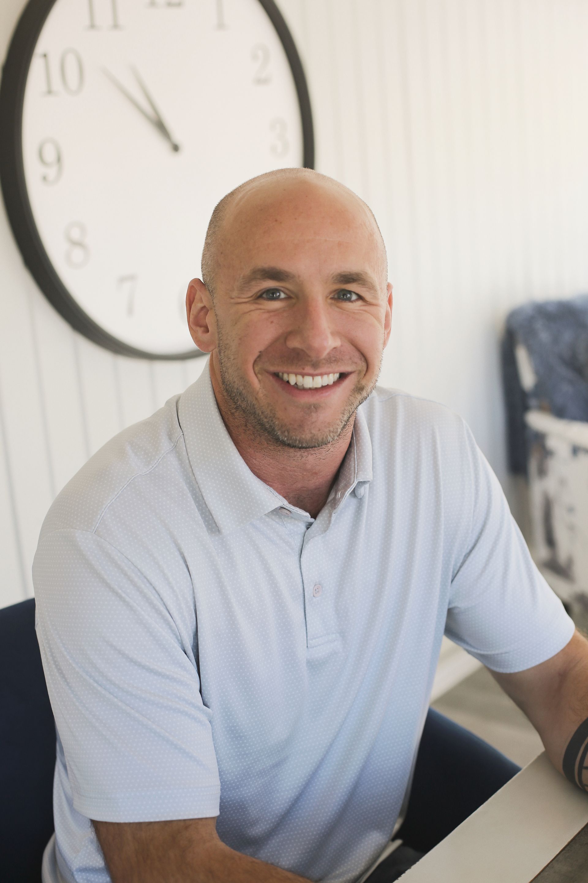 Man with shaved head smiles at the camera, wearing a light blue polo. A clock hangs behind him.