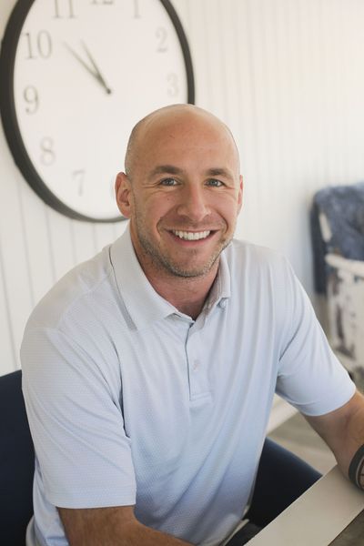 Man with shaved head smiles at the camera, wearing a light blue polo. A clock hangs behind him.