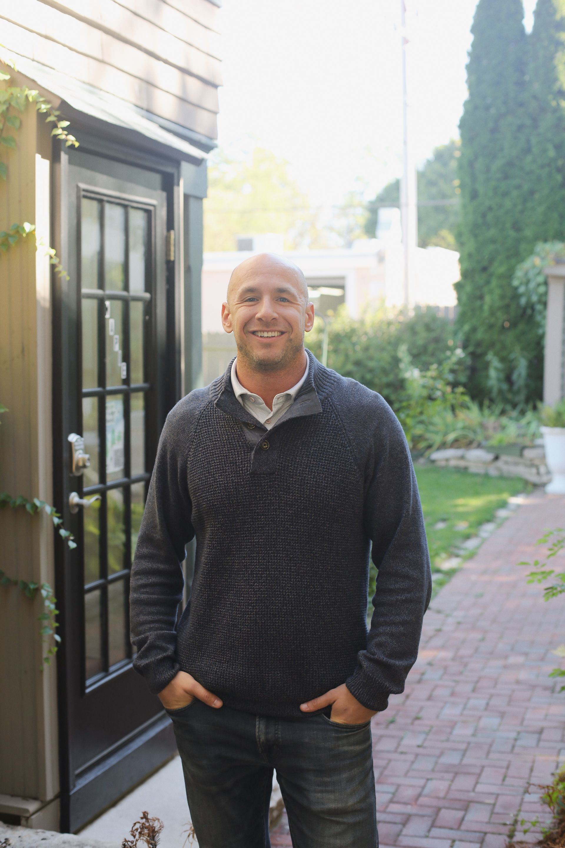 Bald man in a gray sweater smiles, hands in pockets, standing near a dark door and brick walkway.
