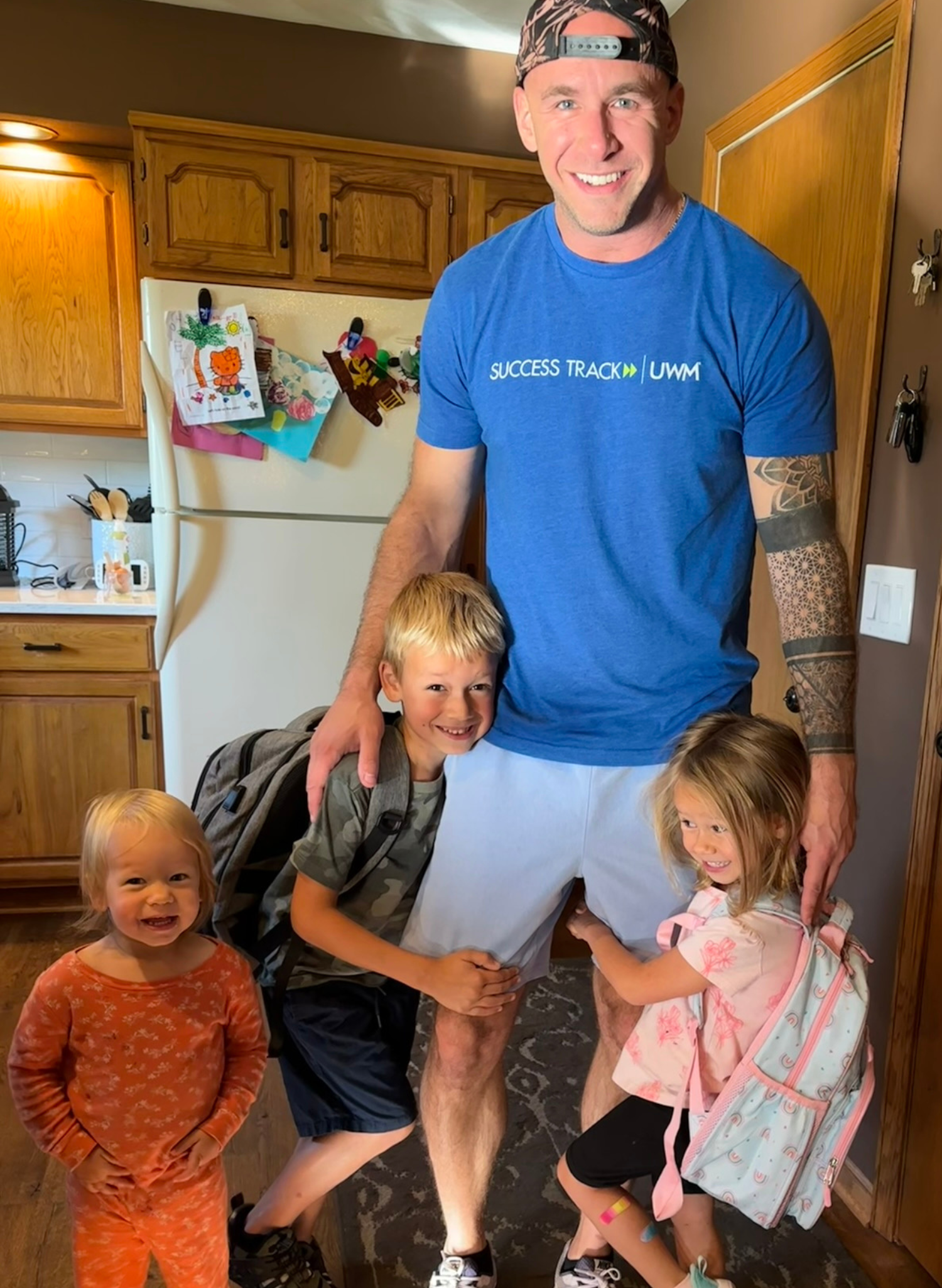 Man smiles with three young children in a kitchen. The children are smiling and the man has a tattoo on his arm.