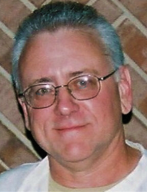 A man wearing glasses and a white shirt is smiling in front of a brick wall.