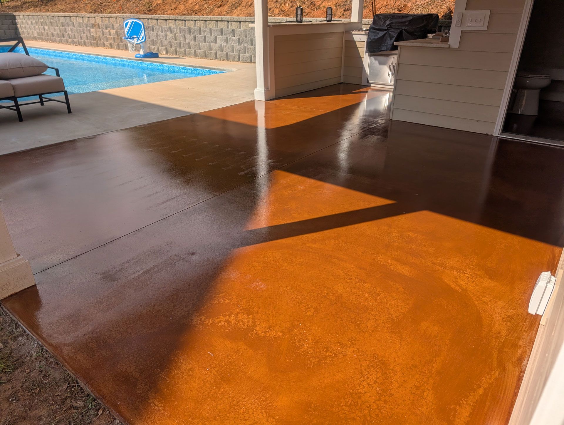 A patio floor with a transition from dark brown to bright orange concrete, adjacent to a pool and a covered outdoor area.