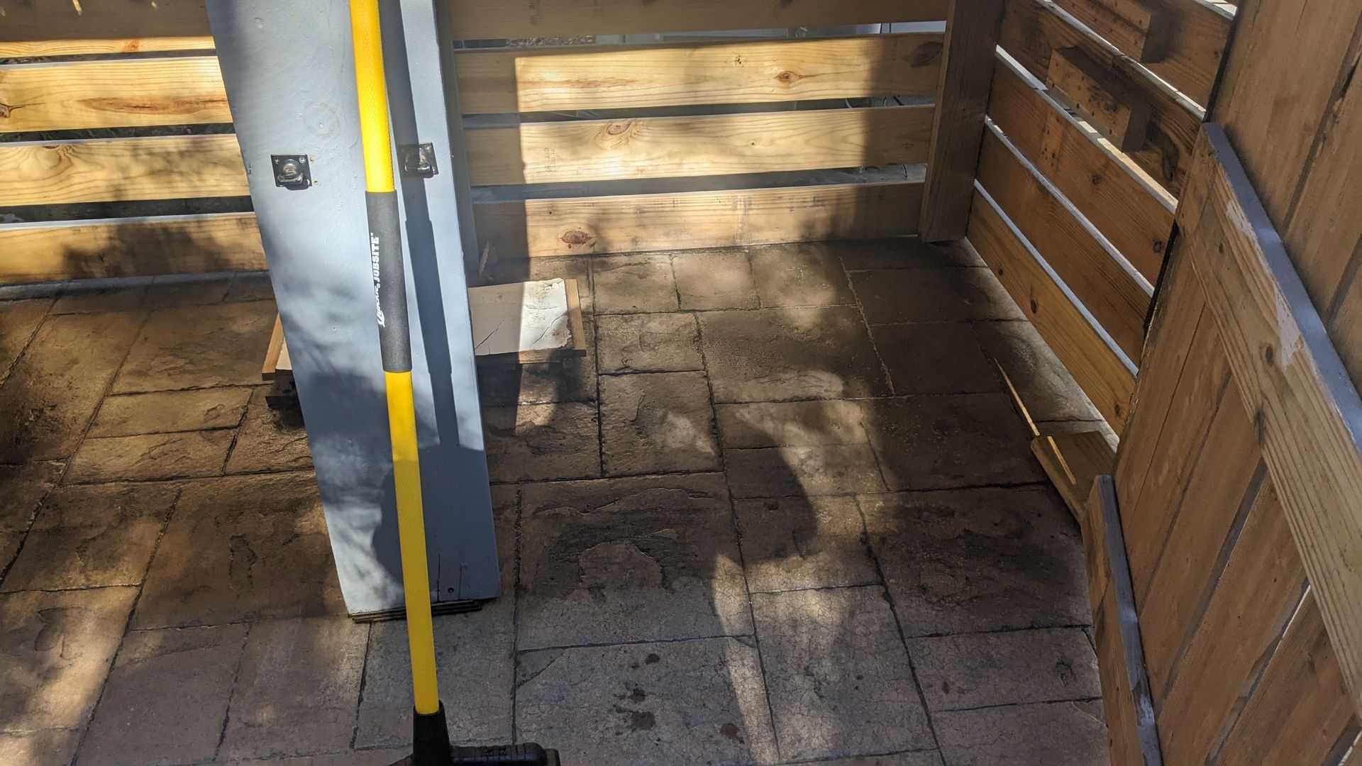 A yellow broom is sitting on a brick floor next to a wooden fence.