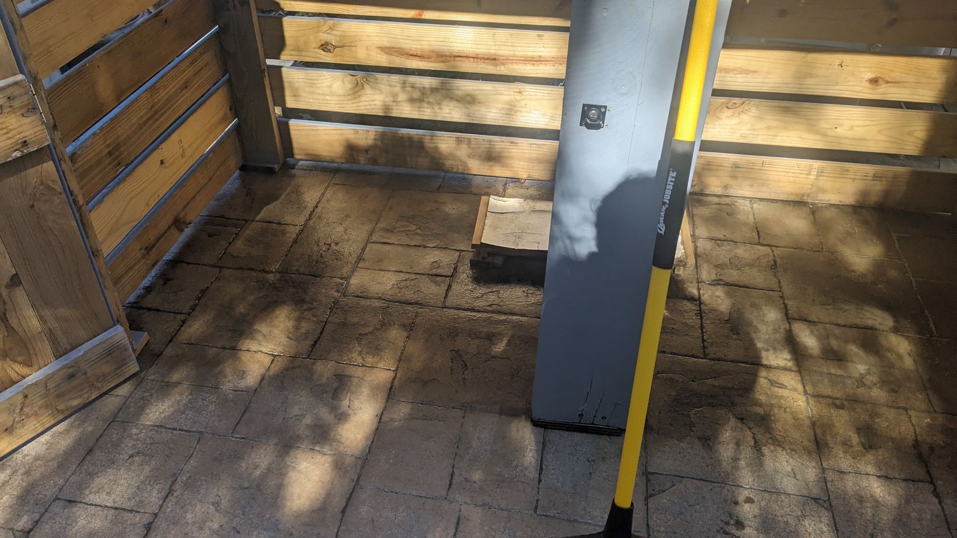 A yellow shovel is sitting on a brick floor next to a wooden fence.