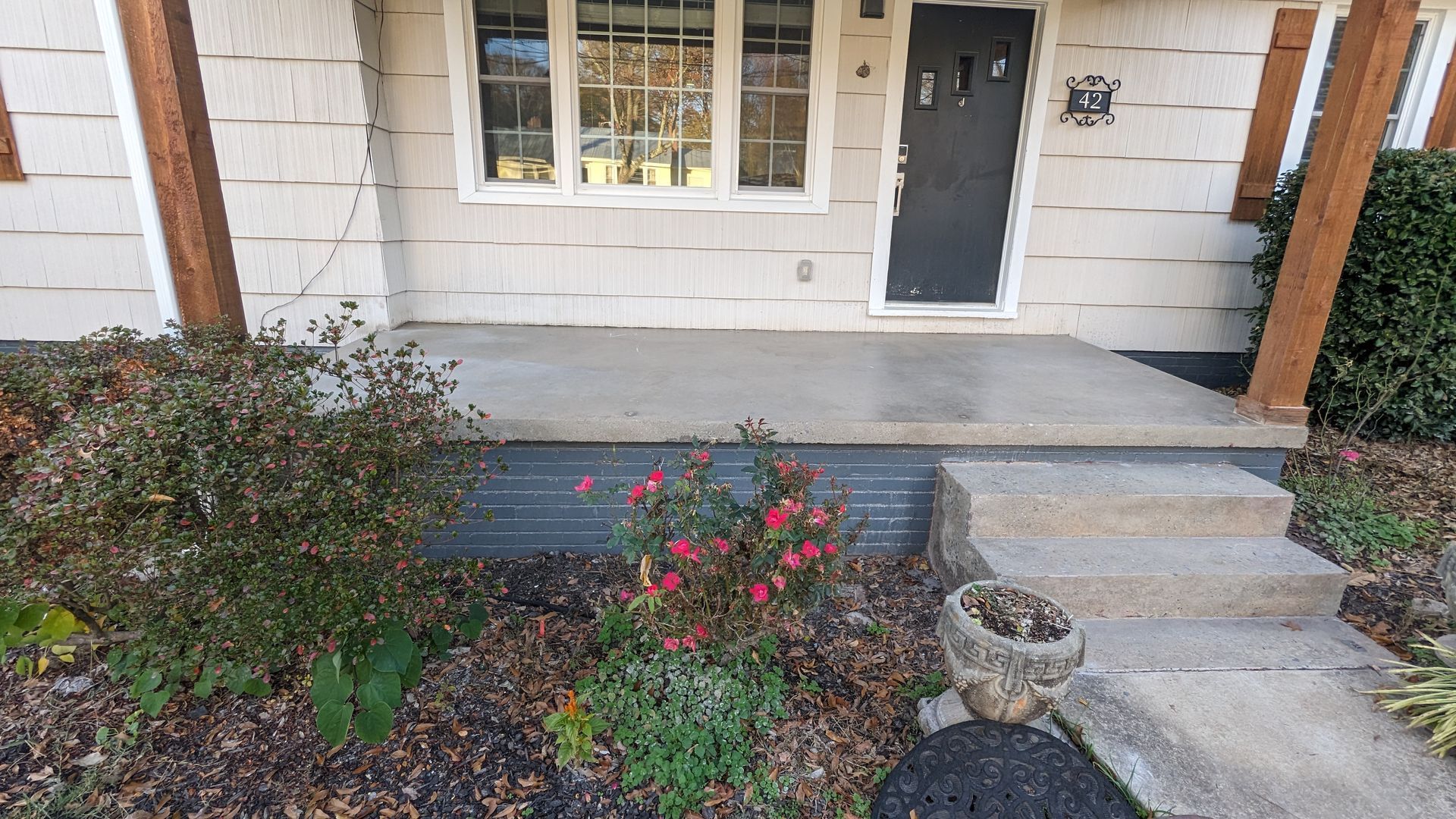A white house with a black door and a porch with flowers in front of it.