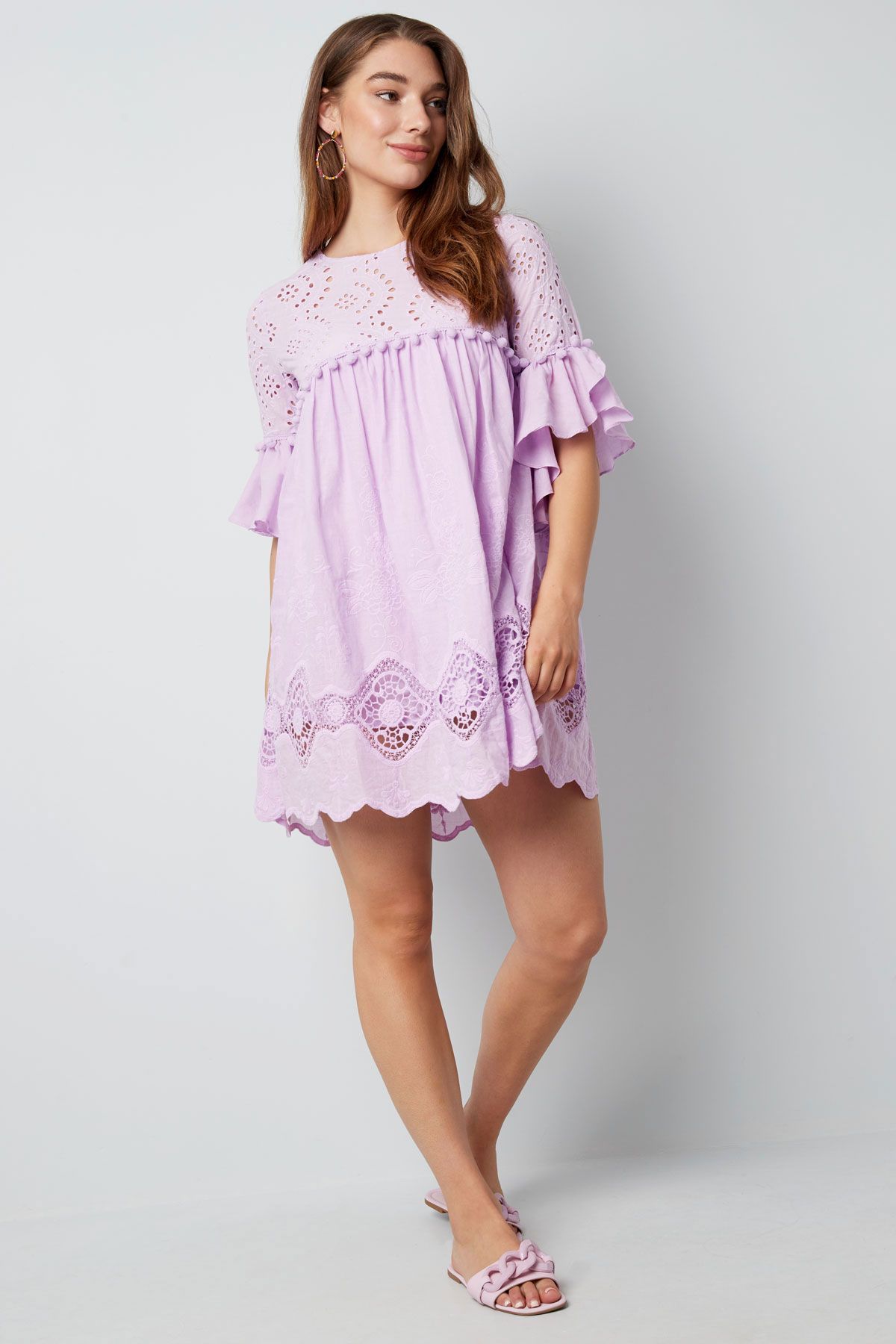 Een model draagt ​​een lavendelkleurige jurk met korte mouwen, ruches en een geschulpte zoom, tegen een grijze achtergrond.