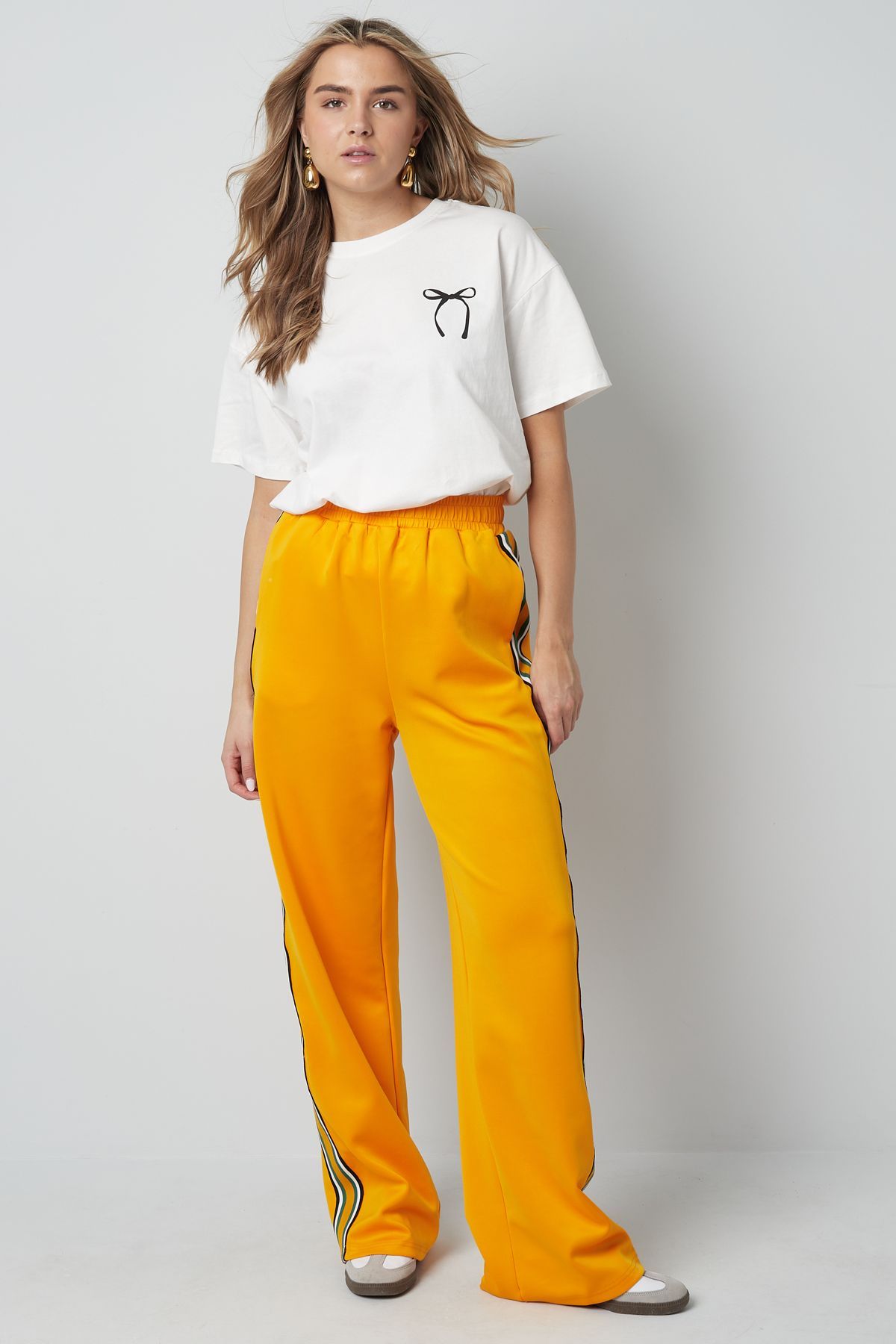 Een persoon draagt ​​een wit T-shirt met een klein logo en een felgele joggingbroek met wijde pijpen en zwarte zijstrepen.