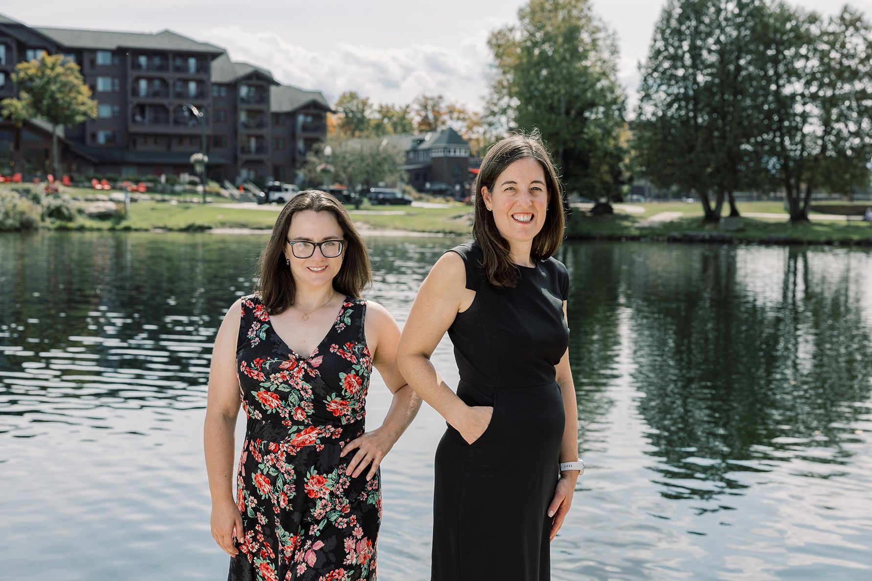 Two  women in formal dresses stand side-by-side on a dock overlooking  lake with a large lodge in the background.