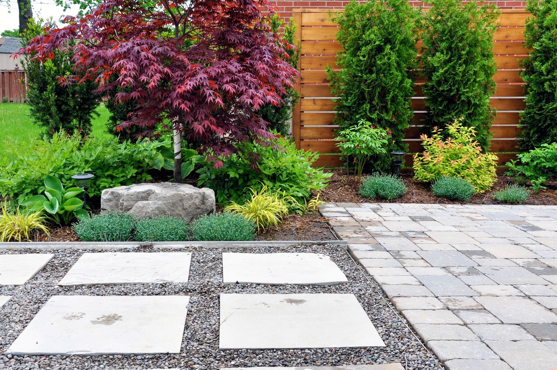 Backyard garden with stone pavers, gravel, shrubs, and a wooden fence