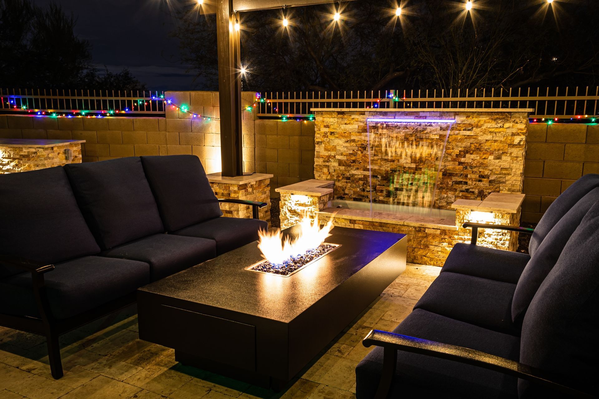 Outdoor patio with blue couches, a fire pit, and a water feature at night. String lights and warm lighting.