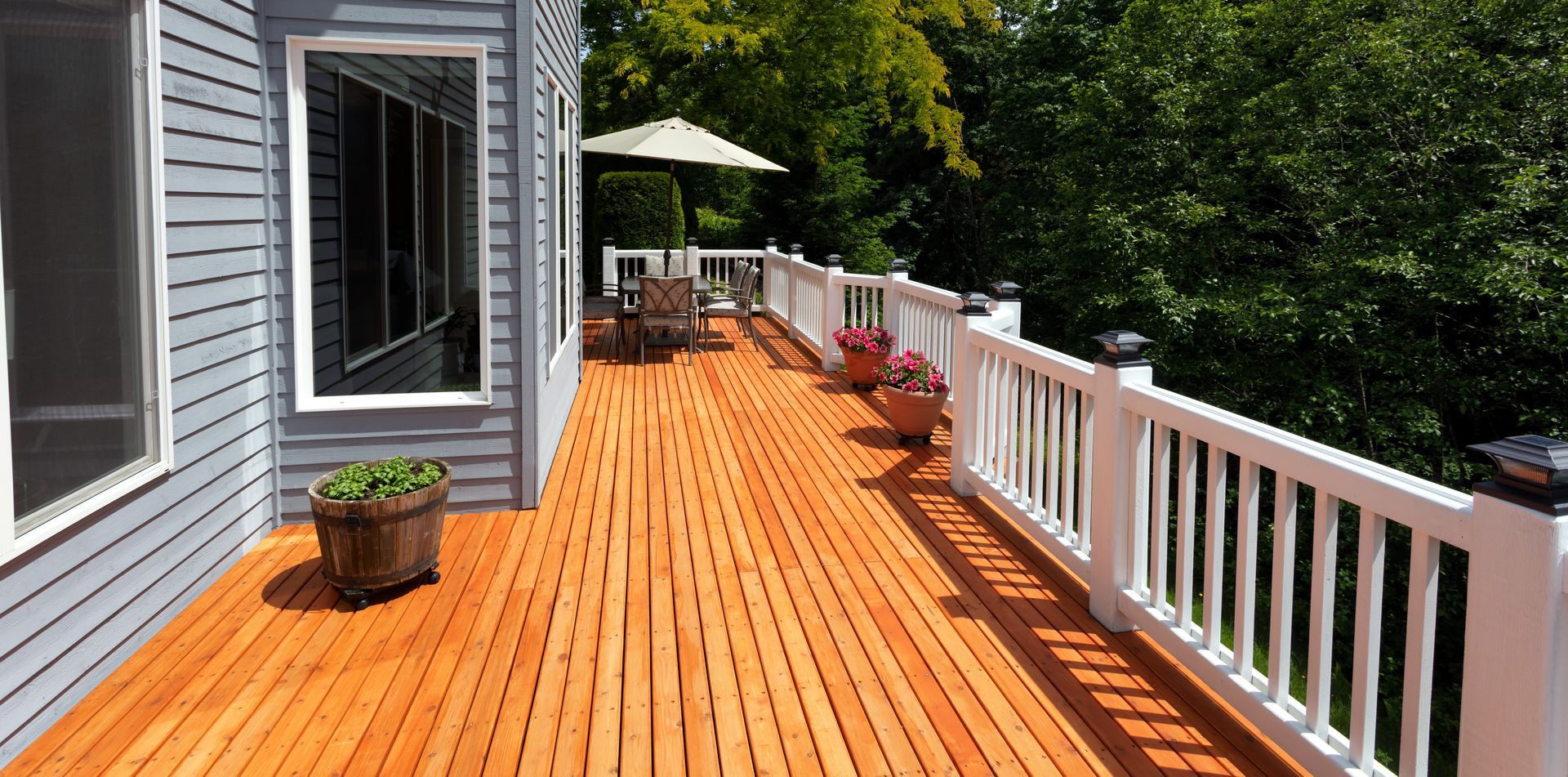 Wooden deck with white railing, potted plants, and an umbrella, beside a blue house, with green trees.