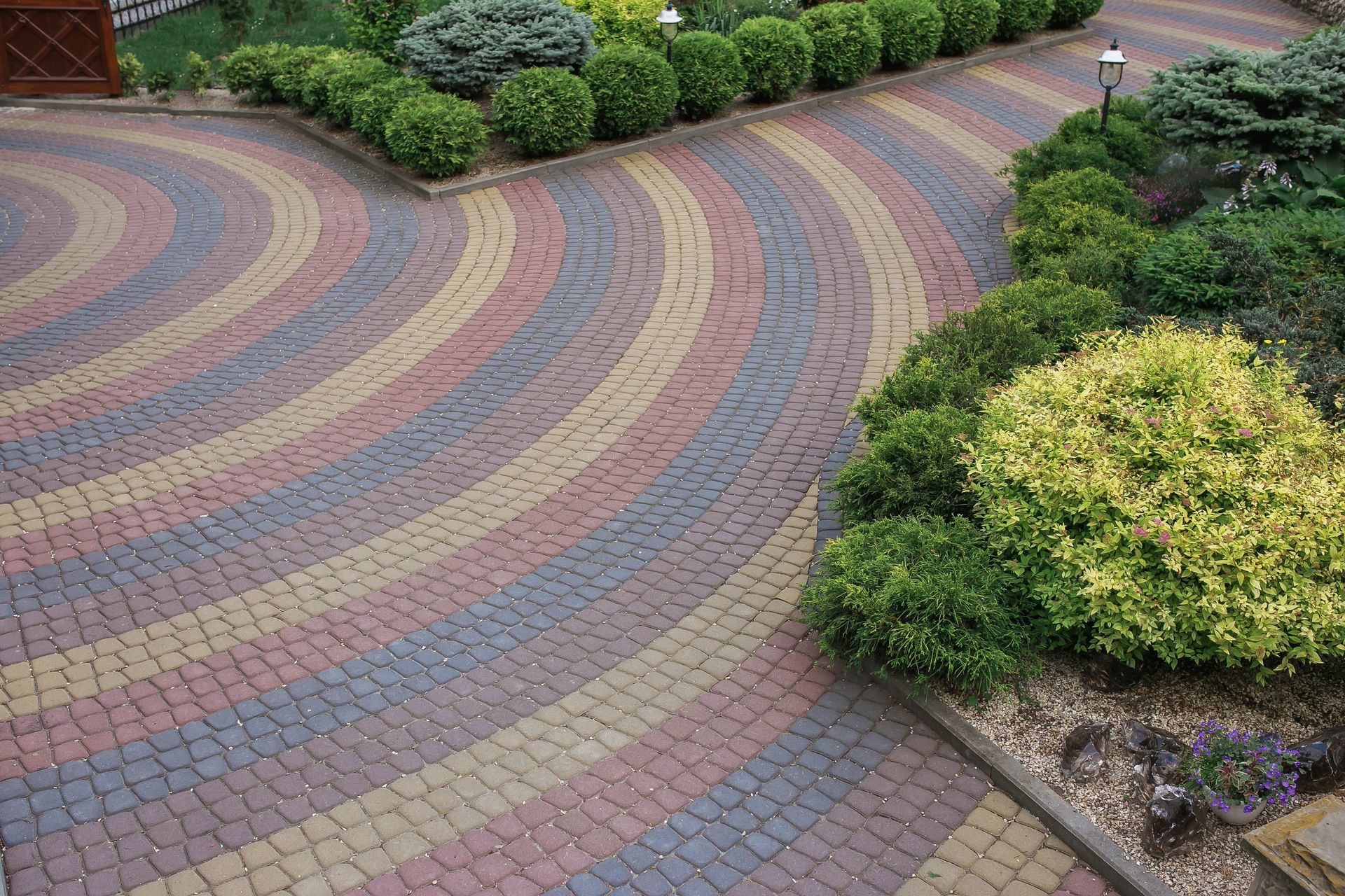 Curved brick driveway with colorful concentric patterns, surrounded by green shrubs and manicured landscaping.