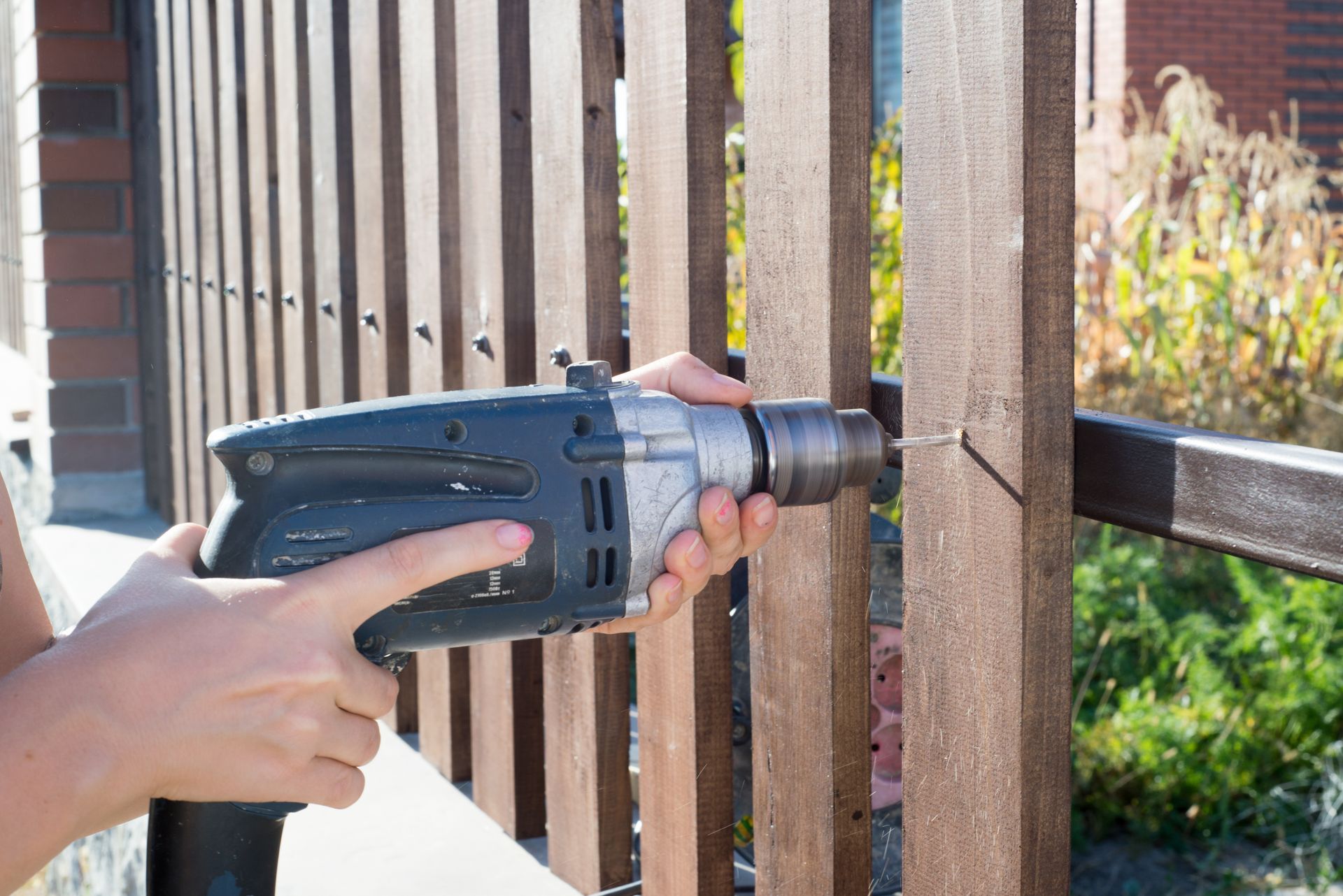 Person using a power drill to fasten a board to a wooden fence. Outdoors, sunny day.