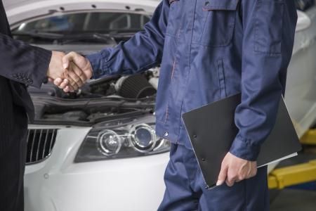 Mechanic in blue coveralls shaking hands with a person in a suit, car with open hood in background.