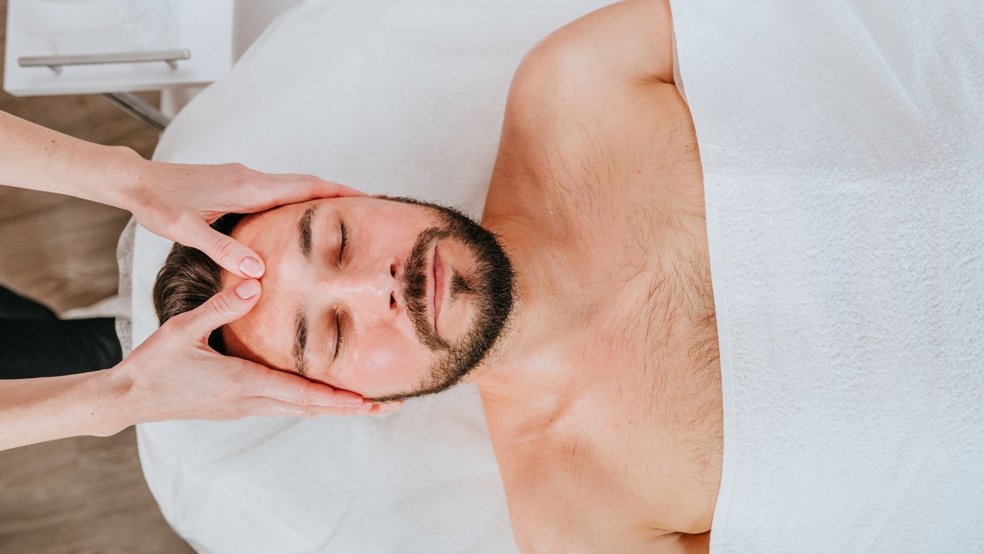 Un homme reçoit un massage du visage, les yeux fermés, sur une table de massage. Des mains massent doucement son front.