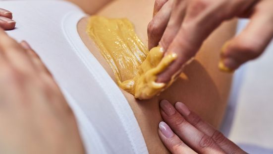 Épilation des aisselles : les aisselles d’une personne sont recouvertes de cire jaune. Une autre personne applique ensuite de la cire supplémentaire.