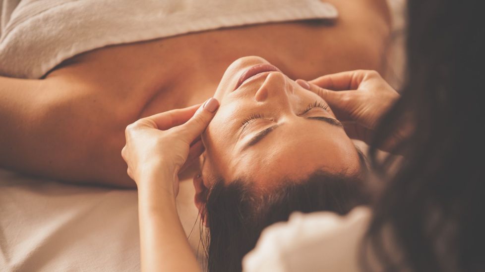 Personne recevant un massage facial ; mains sur les joues ; yeux fermés, serviette blanche.