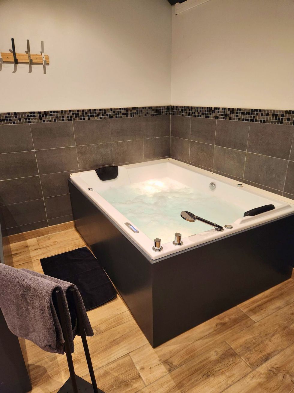 Une baignoire jacuzzi remplie d'eau dans une salle de bains. Mur carrelé gris, sol en bois, serviettes et tapis noirs.