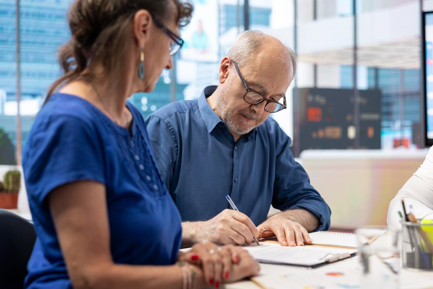 Two people sitting at a desk in a bright office, with one person writing on a document while the other looks on.