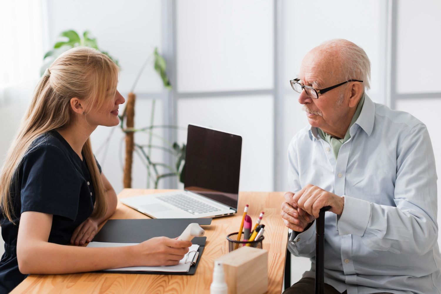 A professional in scrubs consults with an older person holding a cane at a desk with a laptop and documents.