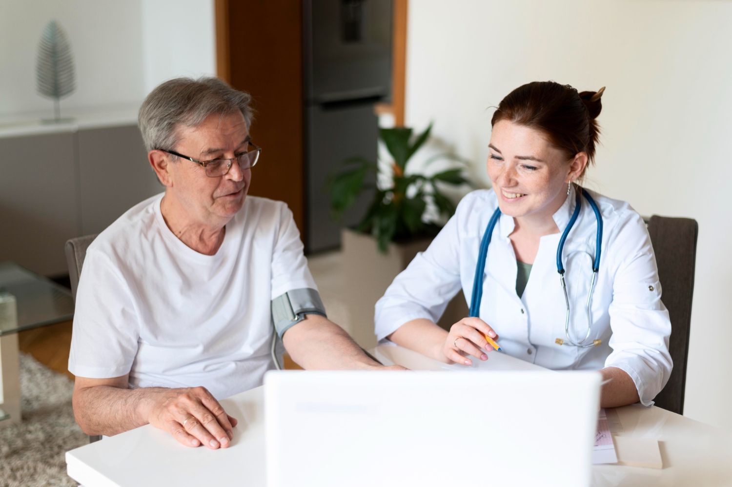 A healthcare worker with a stethoscope reviews health data on a laptop with a patient wearing a blood pressure monitor.