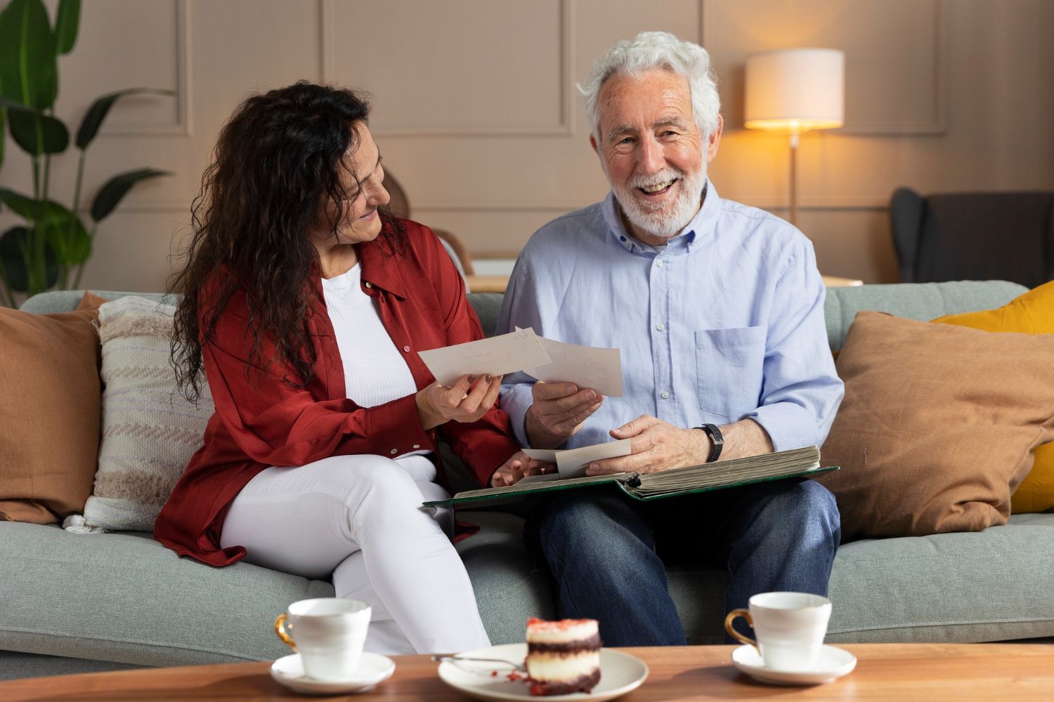 Two people sit on a sofa looking at photos in a book, with coffee and a slice of cake on a table in front of them.