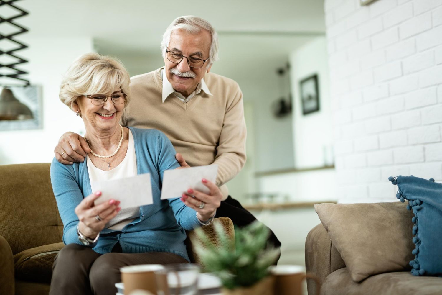 Two people sitting in a cozy living room, smiling while looking at photographs held in their hands.