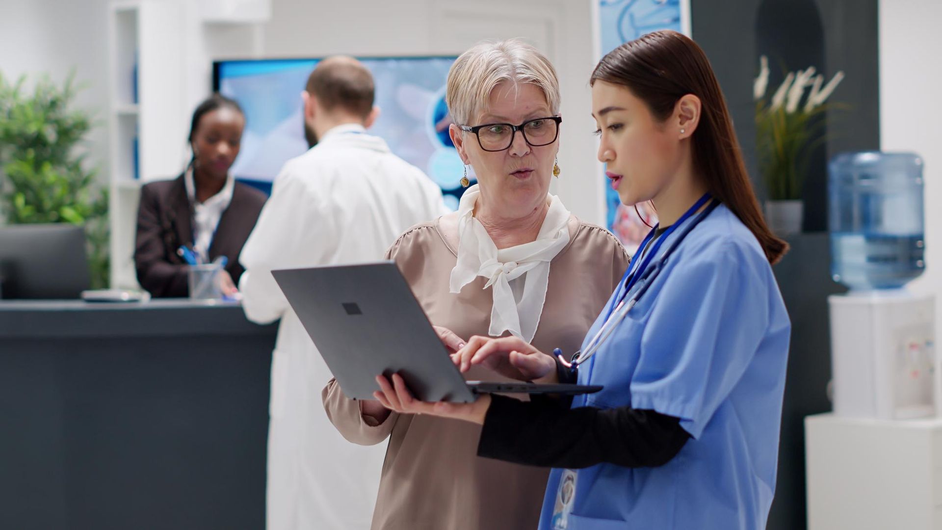 A healthcare worker in scrubs and a person in professional attire review data on a laptop in a medical office.