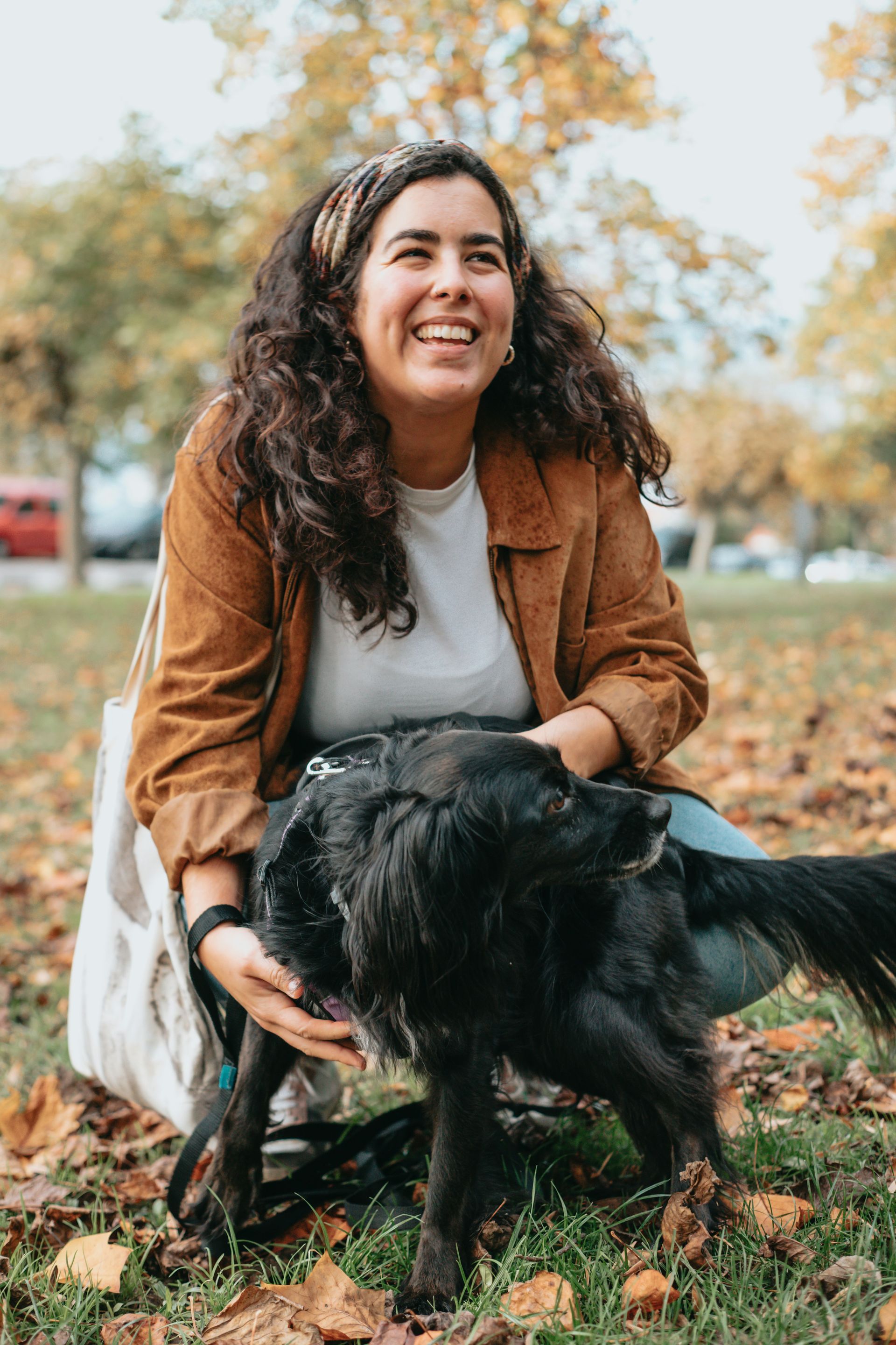A smiling person with long curly hair wearing a brown corduroy jacket crouches in a park, petting a black dog.