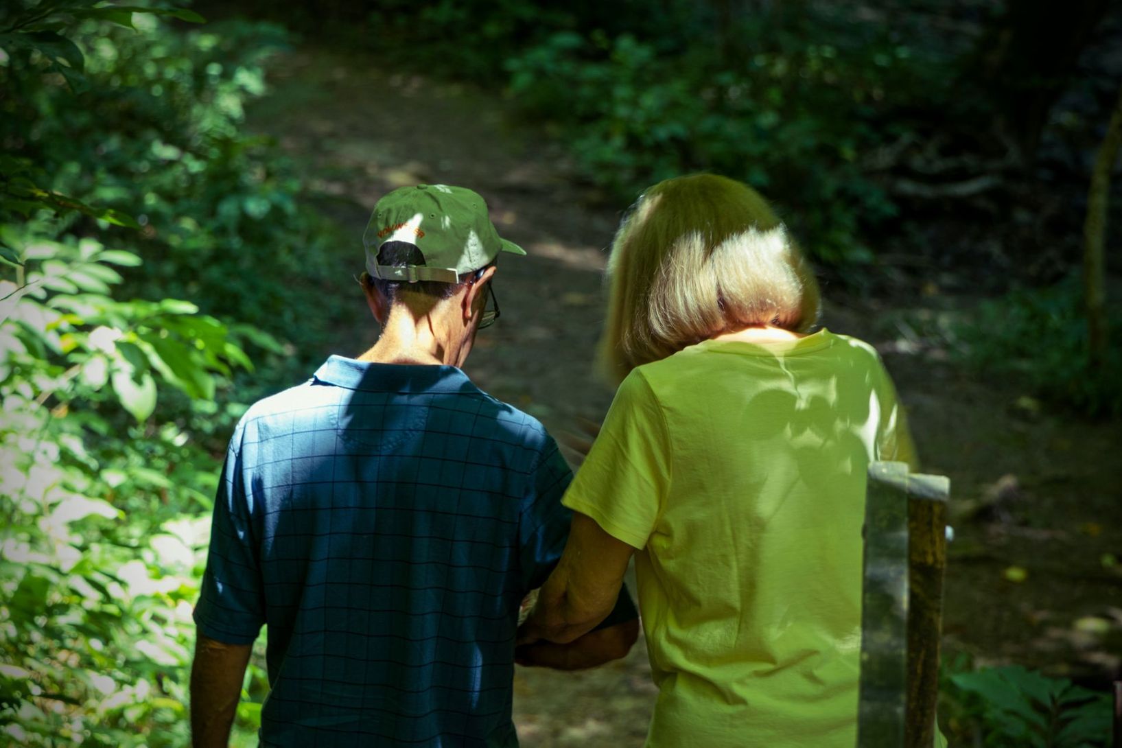 Two people walking together along a shaded forest trail, one wearing a blue shirt and green cap, the other a yellow shirt.