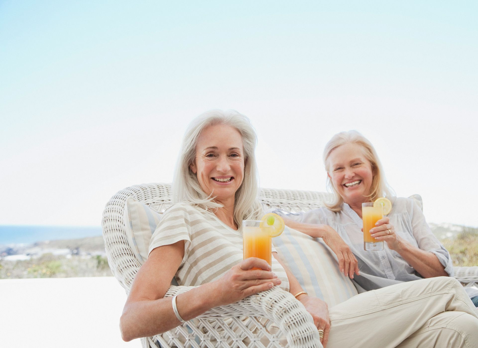 Two women smiling and holding glasses of orange juice while sitting on a wicker sofa outdoors by the sea.