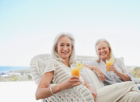 Two women smiling and holding glasses of orange juice while sitting on a wicker sofa outdoors by the sea.