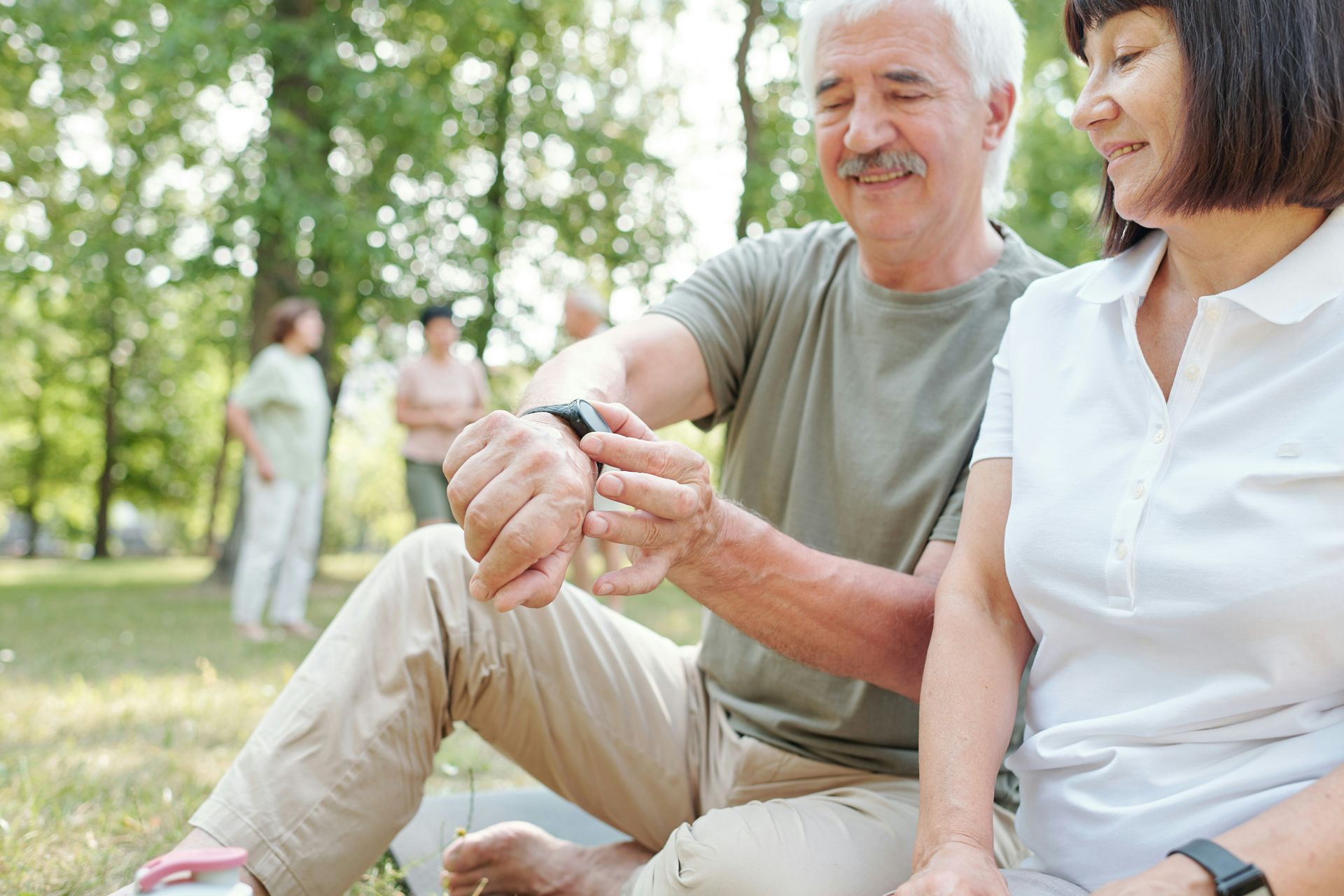 Two people sitting on the grass in a park, one looking at a smartwatch on their wrist while the other watches.