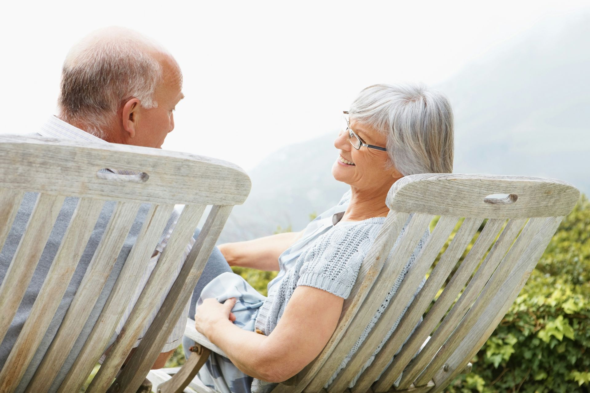 Two people sit in outdoor wooden chairs, facing each other and smiling while engaged in conversation.