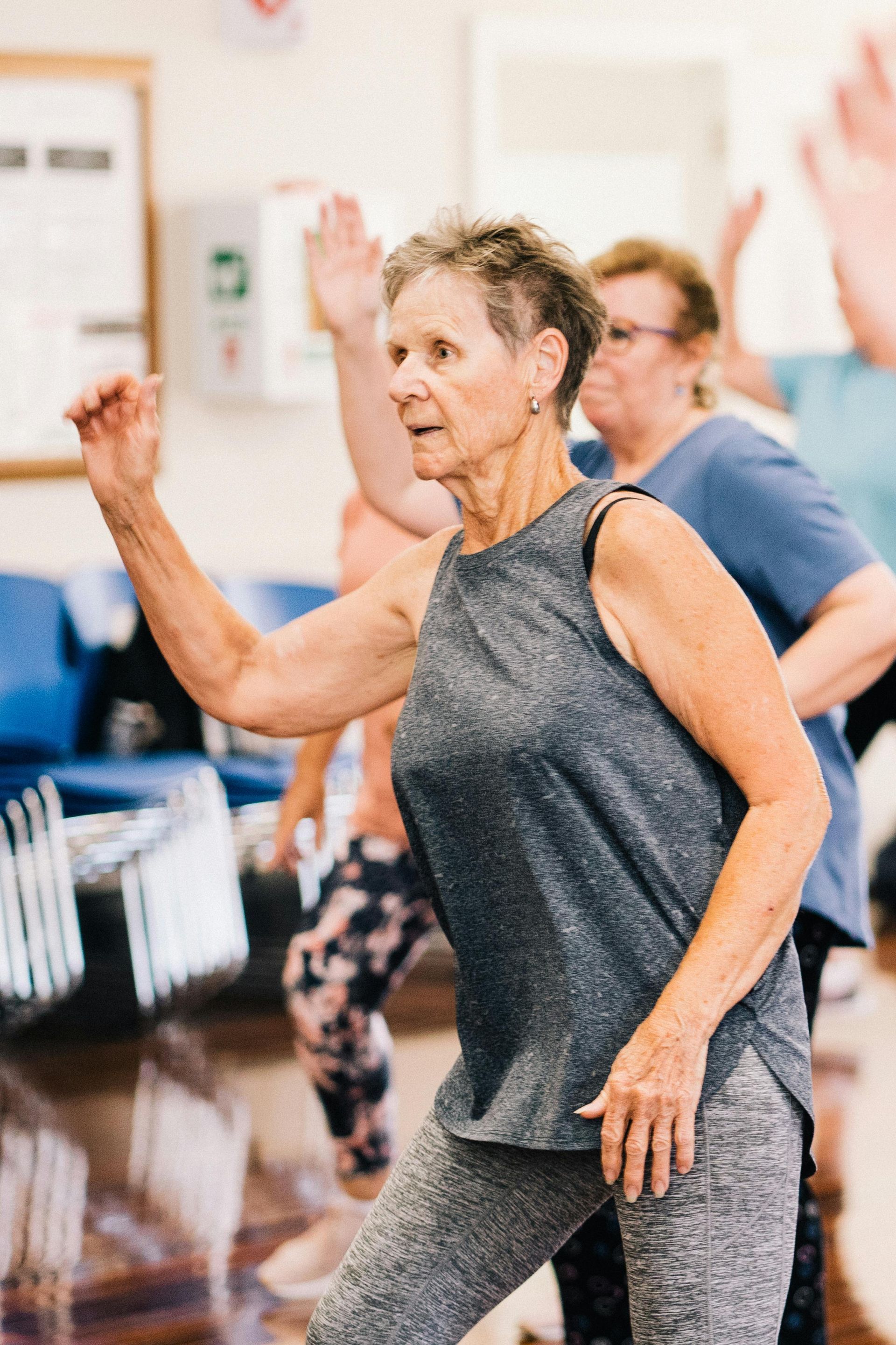 A group of people in an exercise class performing arm movements in a room with blue chairs.