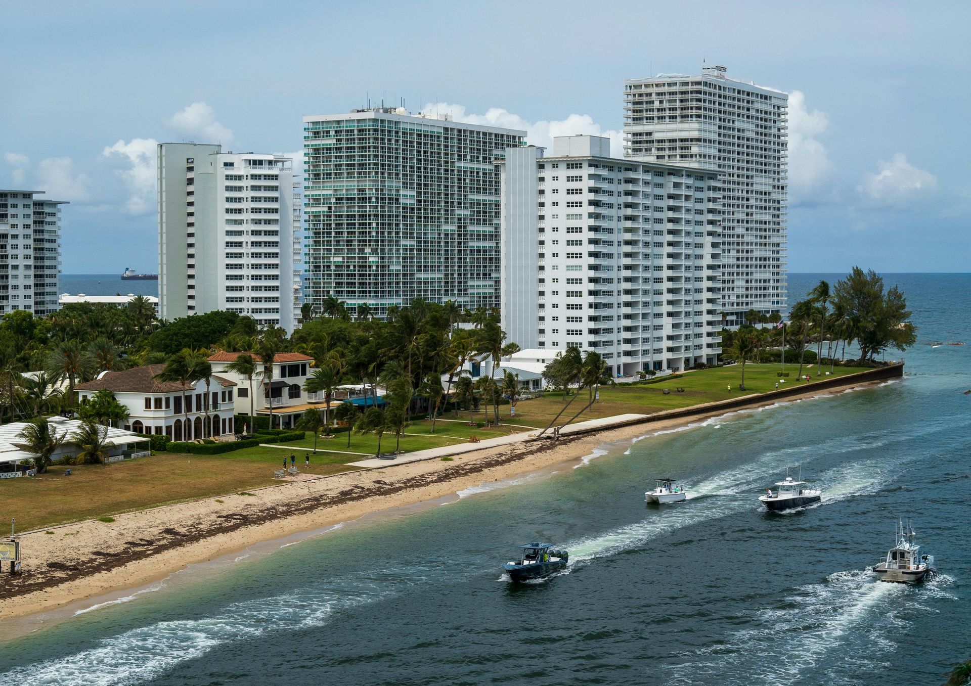 Three boats travel along the coast past a beach lined with tall white apartment buildings under a partly cloudy sky.