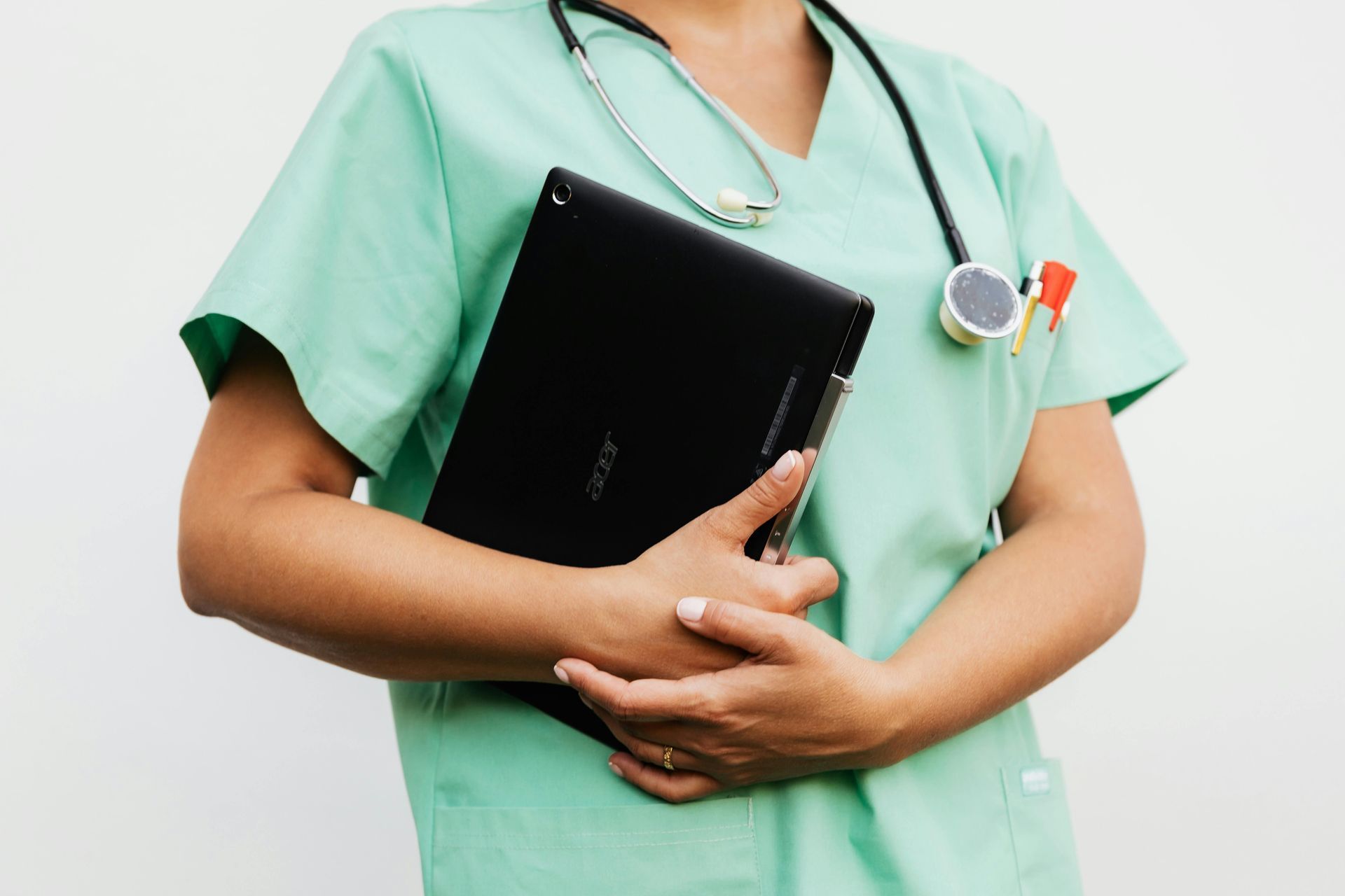 A healthcare professional in mint green scrubs wears a stethoscope while holding a black tablet against their chest.