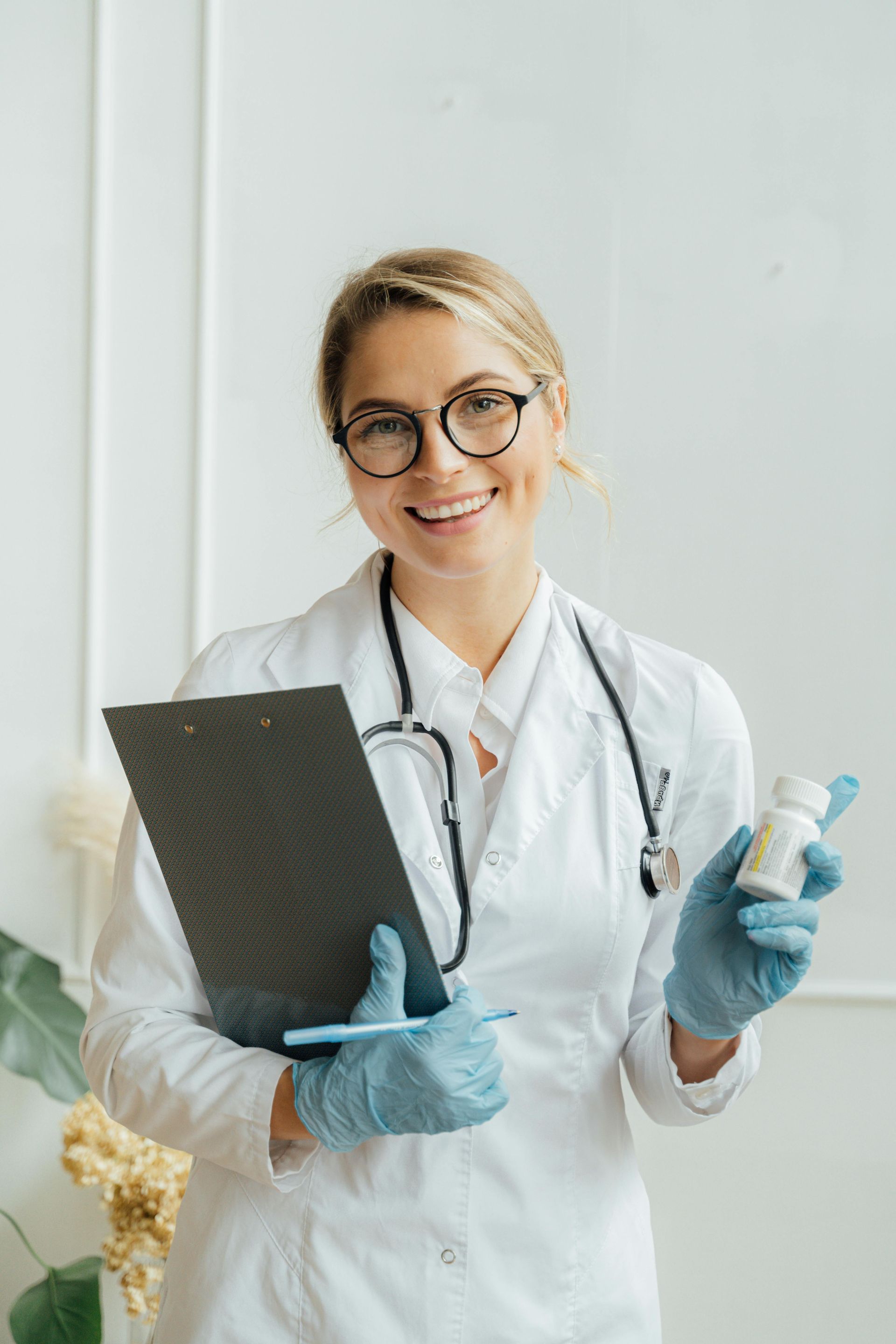 A smiling person in a white lab coat, stethoscope, and blue gloves holds a clipboard and a small pill bottle.