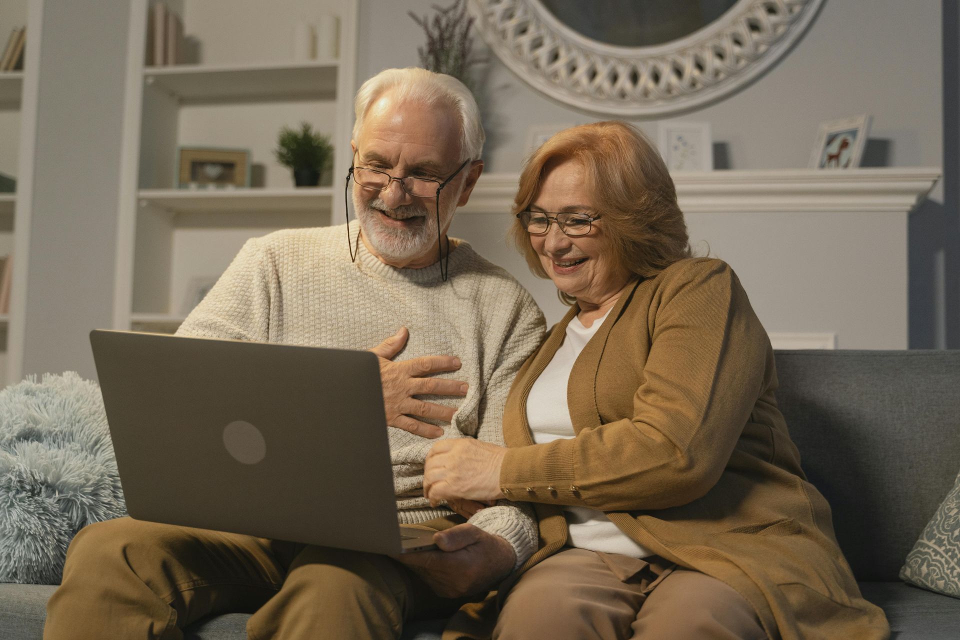 A couple sits on a sofa, smiling while looking at a laptop computer together in a bright, cozy living room.