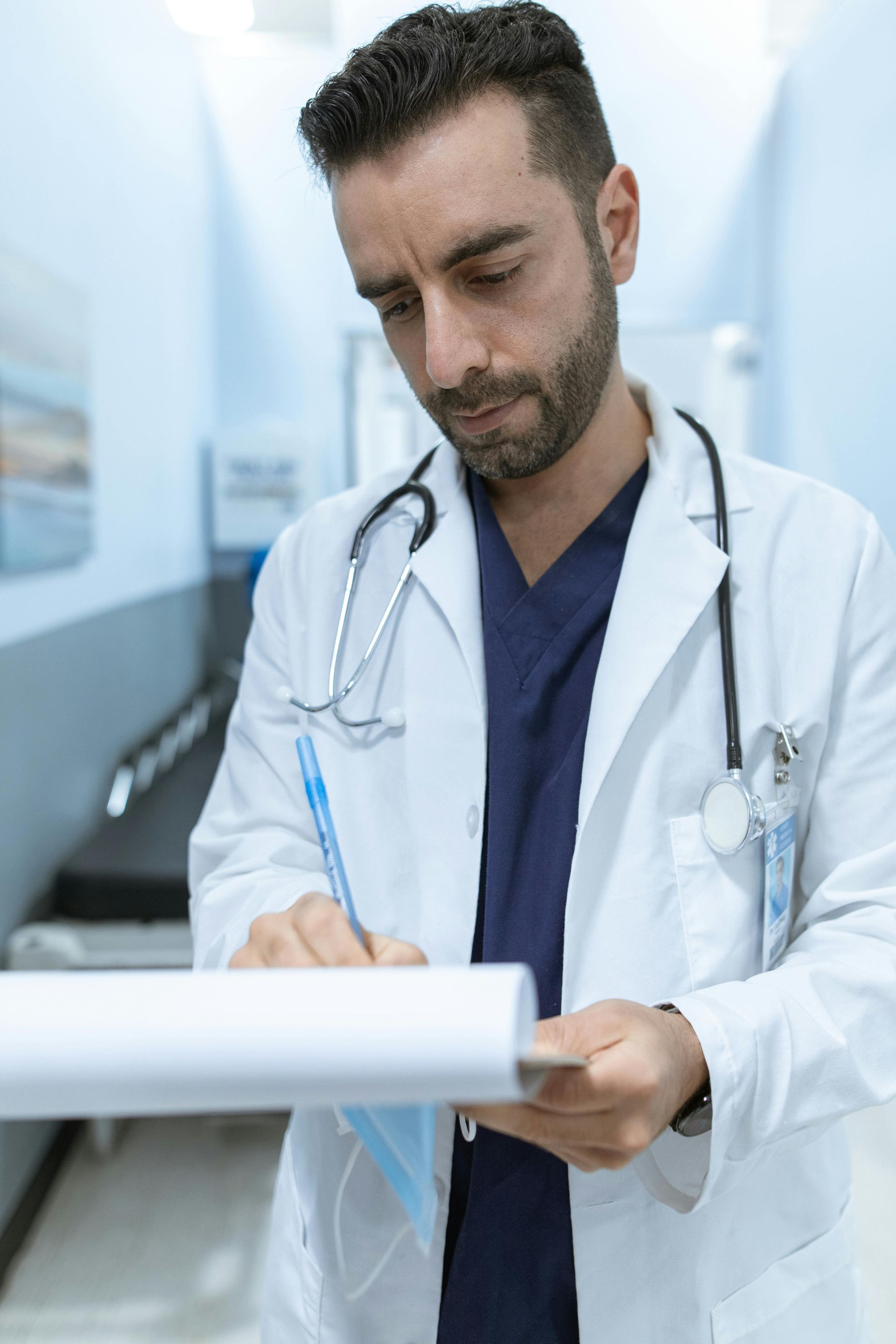 A focused medical professional in a white coat and dark scrubs writes on a clipboard in a hospital hallway.