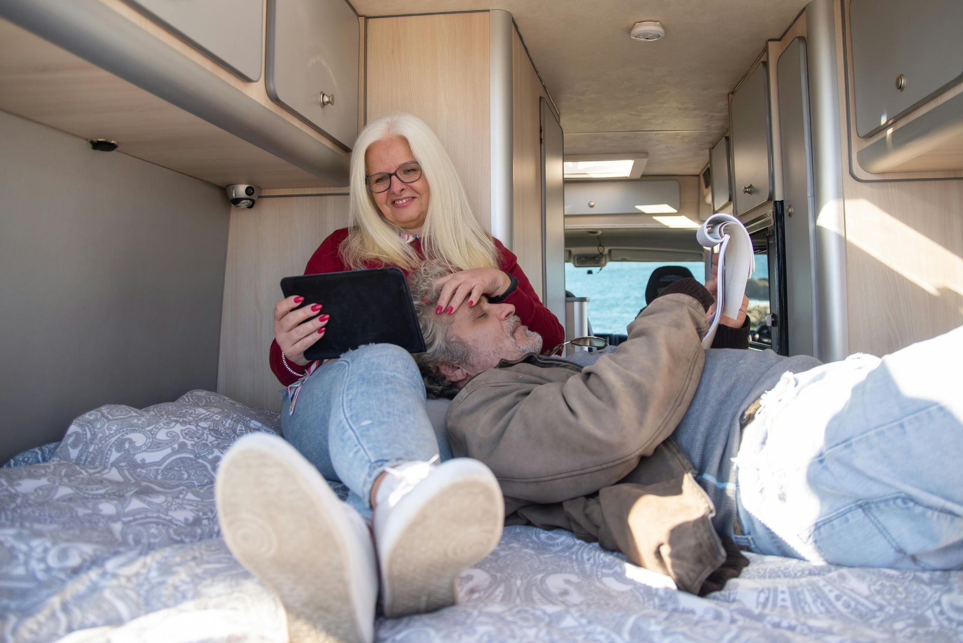 Two people relax on a bed inside a camper van; one sits holding a tablet while the other lies down nearby.